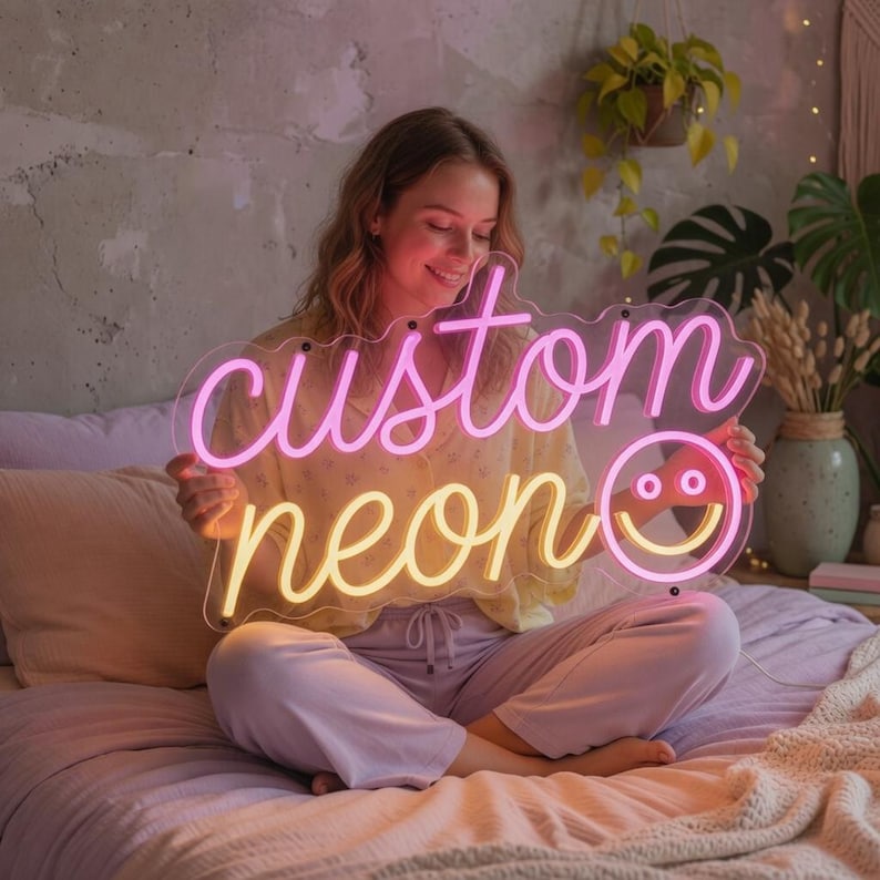 May include: A custom neon sign featuring the word custom in pink script, the word neon in yellow script, and a yellow smiley face icon. The sign is mounted on a clear acrylic backing and emits a bright glow, displaying the text custom neon.