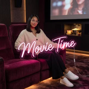 May include: A neon sign with the text Movie Time in a cursive font, emitting a bright pink glow. The sign is constructed with a clear acrylic backing and is connected to a white power cord. The lettering is stylized in a flowing script format.