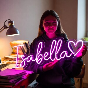 May include: A custom neon sign featuring the name Isabella in a bright pink cursive script, followed by a heart outline. The sign is mounted on a clear acrylic backing and emits a vibrant glow that illuminates the surrounding area in pink light.