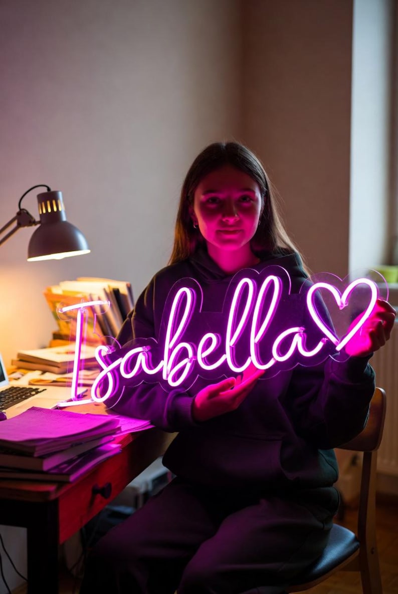 May include: A custom neon sign featuring the name Isabella in a bright pink cursive script, followed by a heart outline. The sign is mounted on a clear acrylic backing and emits a vibrant glow that illuminates the surrounding area in pink light.