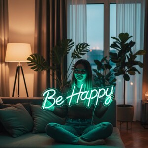 May include: A woman holding a neon sign that reads "Be Happy" in a cursive font. The sign glows with a bright green light. The woman is sitting on a gray couch in a living room with plants and a lamp.