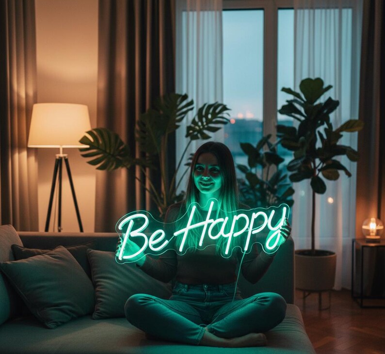 May include: A woman holding a neon sign that reads "Be Happy" in a cursive font. The sign glows with a bright green light. The woman is sitting on a gray couch in a living room with plants and a lamp.