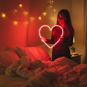 May include: A glowing red neon heart is held in front of a person. The heart is the focal point, with a bright, warm light. The scene is set in a bedroom with a bed and string lights.