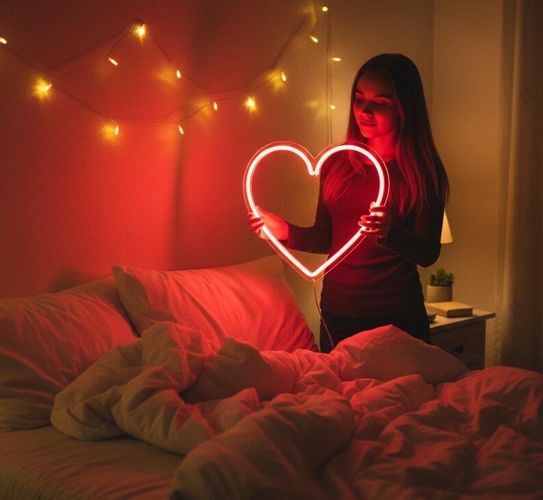 May include: A glowing red neon heart is held in front of a person. The heart is the focal point, with a bright, warm light. The scene is set in a bedroom with a bed and string lights.