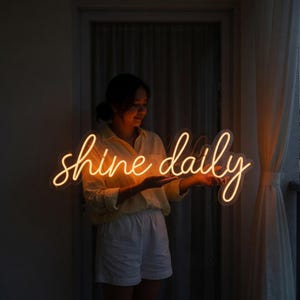 May include: A neon sign featuring the words shine daily in a cursive script font. The sign emits a warm orange glow and is constructed with clear tubing. The text is written in a continuous, flowing style, creating a bright and luminous display.
