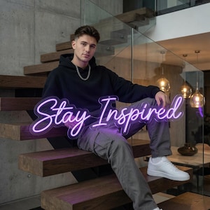 May include: A purple neon sign with the text Stay Inspired in a cursive script font. The sign is held by a person wearing a black hooded sweatshirt, grey cargo pants, and white sneakers, sitting on wooden stairs with a glass railing.