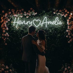 May include: A couple embraces in front of a neon sign that reads "Henry & Amelia" with a heart symbol. The sign is surrounded by greenery and pink flowers, creating a romantic wedding backdrop.