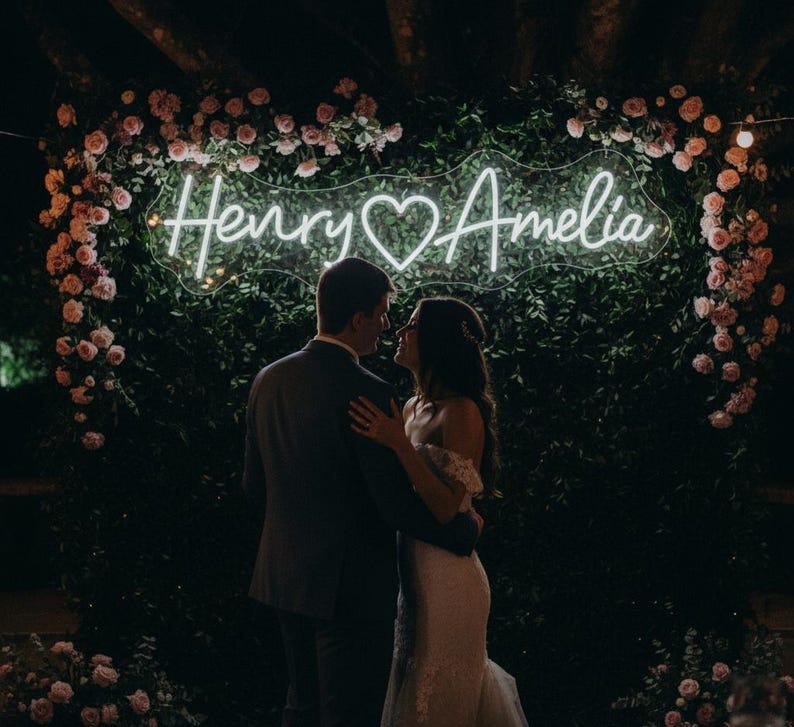 May include: A couple embraces in front of a neon sign that reads "Henry & Amelia" with a heart symbol. The sign is surrounded by greenery and pink flowers, creating a romantic wedding backdrop.