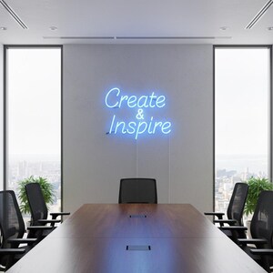 May include: A modern conference room with a large wooden table and black chairs. A neon sign on the wall reads "Create & Inspire" in blue script. Large windows offer a city view, and potted plants add greenery.
