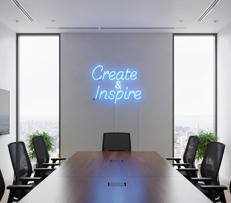May include: A modern conference room with a large wooden table and black chairs. A neon sign on the wall reads "Create & Inspire" in blue script. Large windows offer a city view, and potted plants add greenery.
