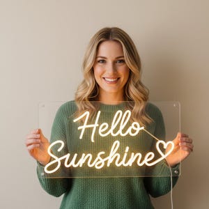 May include: A woman holding a clear acrylic sign with the words "Hello Sunshine" in a warm, glowing neon light. The sign has a heart symbol at the end. The woman is wearing a green sweater.