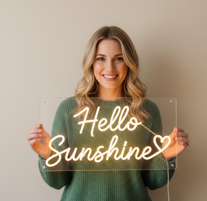 May include: A woman holding a clear acrylic sign with the words "Hello Sunshine" in a warm, glowing neon light. The sign has a heart symbol at the end. The woman is wearing a green sweater.