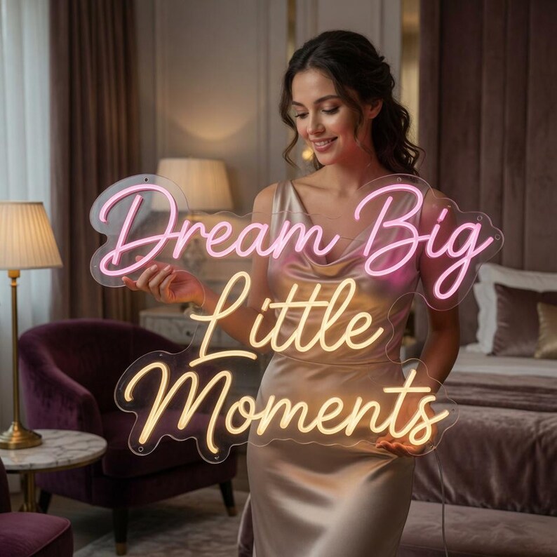 May include: A clear acrylic neon sign featuring the text "Dream Big Little Moments" in a cursive font. The word "Dream" is in pink, while "Big Little Moments" is in a warm yellow hue. The sign has a contoured edge and mounting holes.
