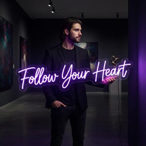 May include: A bright purple neon sign featuring the cursive text Follow Your Heart. The sign is constructed with a clear acrylic backing and is illuminated, casting a vibrant glow. It is held horizontally, displaying the script lettering clearly.