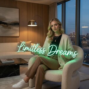 May include: A neon sign featuring the text "Limitless Dreams" in a glowing light green script font. The sign is constructed with a clear acrylic backing and is held by a person wearing a cream-colored cardigan, a black top, and tan trousers.