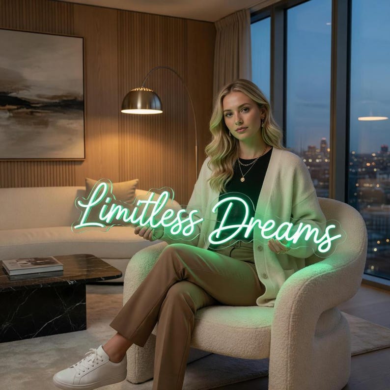 May include: A neon sign featuring the text "Limitless Dreams" in a glowing light green script font. The sign is constructed with a clear acrylic backing and is held by a person wearing a cream-colored cardigan, a black top, and tan trousers.