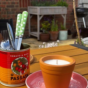May include: A white candle in a terracotta pot sits on a pink patterned plate next to a metal tomato and basil passata can filled with cutlery featuring green handles with white polka dots. The items are arranged on a wooden surface.