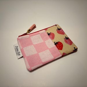 May include: A small, rectangular coin purse with a pink and white checkered pattern on one side and a strawberry print on the other. The purse has a red zipper and a small pink tassel. The tag reads "Polka Dot Threads".