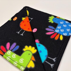 May include: A black fabric coaster with a colorful chicken design. The chicken is depicted with a bright green body, yellow beak, and rainbow-colored feathers.
