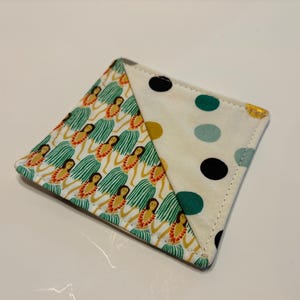 May include: A square fabric coaster with a white background and two different patterns. One side features a repeating pattern of hula dancers in green skirts and orange leis. The other side features a polka dot pattern with black, teal, and gold dots.