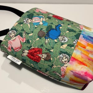 May include: A crossbody bag featuring a green fabric with a repeating pattern of four women in various colored outfits amidst large green leaves. The bag has a black strap and a side panel with a multicolored watercolor-style print. A white tag reads Polka Dot Threads.