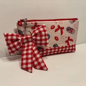 May include: A small, zippered pouch with a red and white gingham bow keychain. The pouch is decorated with red bows, hearts, and lipstick prints on a light pink background. The bottom trim is red and white gingham.
