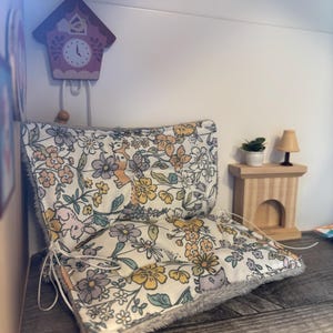 May include: A miniature dollhouse scene featuring a floral-patterned cushion with a gray fur trim. A small wooden cuckoo clock and a miniature wooden table with a lamp and a plant are also visible. The color palette includes soft pastels and natural wood tones.