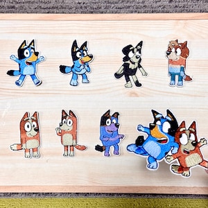 Puede incluir: Una colección de parches bordados con personajes de perros de dibujos animados en varias poses. Los parches muestran una gama de colores, incluyendo azul, marrón y negro, con expresiones faciales y contornos detallados. Los parches están dispuestos sobre una superficie de madera clara.