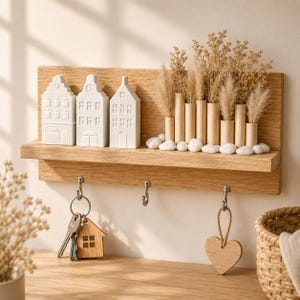 Peut inclure: Une étagère murale en bois de chêne avec trois ornements en céramique blanche en forme de maison, une rangée de vases cylindriques en carton avec des herbes séchées et des galets blancs. Trois crochets métalliques portent un porte-clés et un cœur en bois.