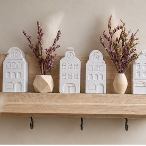 Peut inclure: Une étagère murale en bois clair avec trois crochets en métal, présentant trois ornements blancs en forme de maison et deux petits vases en bois contenant des fleurs séchées violettes et blanches disposés sur l'étagère.