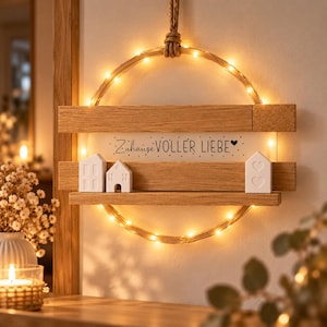 Könnte beinhalten: Ein kreisförmiger Wandbehang aus Holz mit einer Lichterkette aus warmen LEDs, zwei horizontalen Holzleisten und einer weißen rechteckigen Platte mit dem schwarzen Text "Zuhause VOLLER LIEBE" und einem kleinen Herz. Drei weiße Keramikhausfiguren.