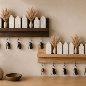 Peut inclure: Deux étagères murales en bois avec des crochets pour clés. L'étagère supérieure est en bois foncé avec des ornements blancs en forme de maison et des herbes séchées. L'étagère inférieure est en bois clair avec des ornements similaires. Clés incluses.