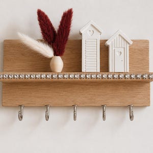 Peut inclure: Un porte-clés mural en bois clair rectangulaire avec cinq crochets en métal argenté. L'étagère présente une rangée de cristaux transparents incrustés. Sur l'étagère se trouvent un petit vase en bois clair avec des herbes séchées bordeaux et blanches.