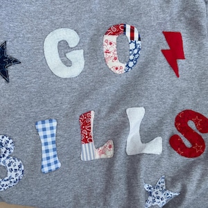 May include: A heather gray sweatshirt featuring fabric applique letters and shapes. The text reads GO BILLS. The letters and shapes are crafted from various patterned fabrics including blue stars, white, red floral, blue gingham, red paisley, and red lightning bolt.
