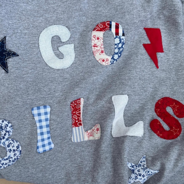 May include: A heather gray sweatshirt featuring fabric applique letters and shapes. The text reads GO BILLS. The letters and shapes are crafted from various patterned fabrics including blue stars, white, red floral, blue gingham, red paisley, and red lightning bolt.
