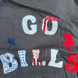 May include: A dark gray long-sleeve shirt featuring fabric applique letters that spell GO BILLS. The letters are crafted from various patterned fabrics including blue gingham, red and white stripes, and floral prints, accompanied by a blue star and red lightning bolt.