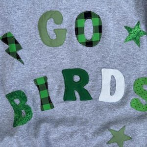 May include: A heather gray sweatshirt featuring fabric applique letters that spell out GO BIRDS. The letters are crafted in various shades of green, including buffalo plaid, solid, and polka dot patterns, accented by green fabric stars and a lightning bolt.