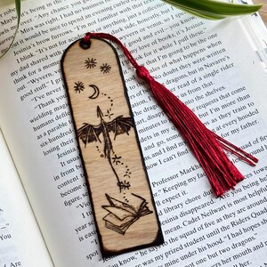 May include: Wooden bookmark with a dragon design, stars, and a crescent moon. The bookmark has a red tassel and is placed on top of an open book. The dragon is flying towards an open book.
