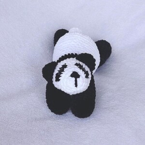 May include: A handmade crochet panda bear toy in black and white. The panda has black ears, eyes, and limbs, with a white head and body. The toy is lying on its back, showcasing its soft, textured design.