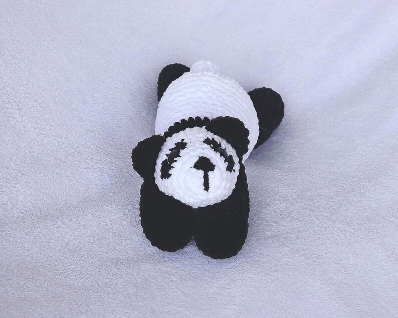 May include: A handmade crochet panda bear toy in black and white. The panda has black ears, eyes, and limbs, with a white head and body. The toy is lying on its back, showcasing its soft, textured design.