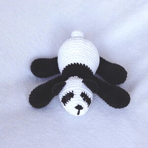 May include: A handmade crochet panda bear in black and white. The panda has a white head and body with black arms, legs, and ears. The face features black eyes and a small black nose. The panda is lying down.