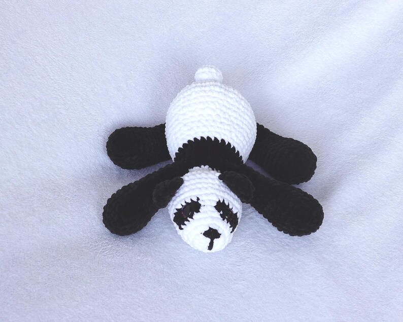May include: A handmade crochet panda bear in black and white. The panda has a white head and body with black arms, legs, and ears. The face features black eyes and a small black nose. The panda is lying down.