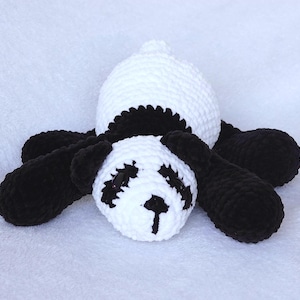 Può includere: Un giocattolo fatto a mano all'uncinetto a forma di panda bianco e nero. Il panda ha la testa e il corpo bianchi con dettagli neri intorno agli occhi, alle orecchie e agli arti. Il giocattolo è sdraiato su una superficie bianca.