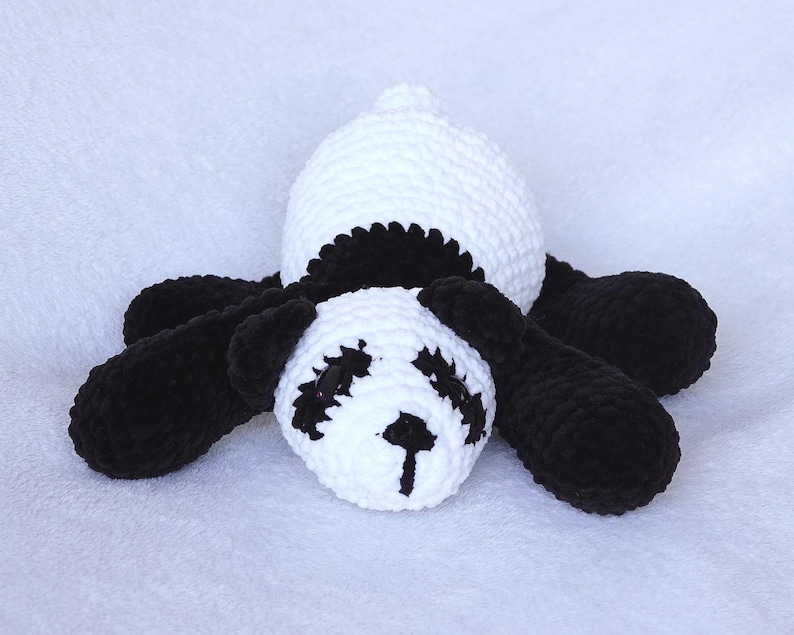 May include: A handmade crochet panda bear toy in black and white. The panda has a white head and body with black accents around the eyes, ears, and limbs. The toy is lying down on a white surface.