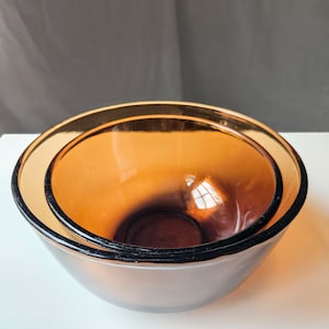 May include: Two amber-coloured glass mixing bowls nested together. The bowls have a rounded shape with a slightly flared rim. The glass has a warm, translucent quality, and the rims are outlined with a dark colour.