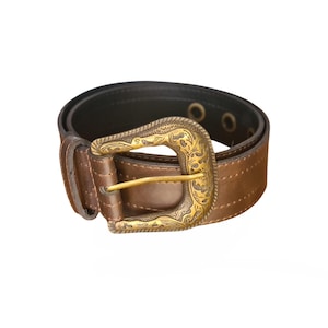 Op de afbeelding: Een bruine leren riem met een decoratieve goudkleurige gesp. De gesp heeft een sierlijk bloemmotief. De riem heeft een donkerbruine binnenkant en is ontworpen met een reeks oogjes.