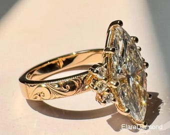 May include: A gold ring with a large, marquise-cut diamond as the centerpiece. The band features intricate, engraved detailing. Small accent diamonds are set around the main stone. The ring is displayed on a light-colored surface, with the brand name "ElaraDiamond" visible.