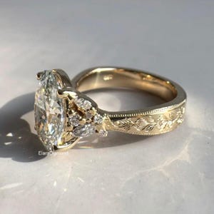 May include: A gold ring with a large, clear, oval-cut gemstone as the centerpiece. The ring features smaller accent stones and floral engravings on the band. The ring is set on a white surface and is well-lit, highlighting its details.