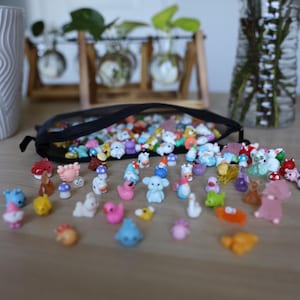 May include: A collection of miniature figurines, including animals, mushrooms, and gnomes, spilling from a black mesh bag onto a wooden surface. The figurines are brightly colored, with details like ducks, bears, and other creatures.