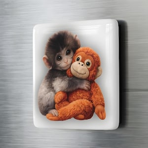 May include: A rectangular magnet featuring two cartoon monkeys. One monkey, with brown and gray fur, hugs a plush orange monkey. The image has a soft, illustrative style, with a white background and rounded corners.